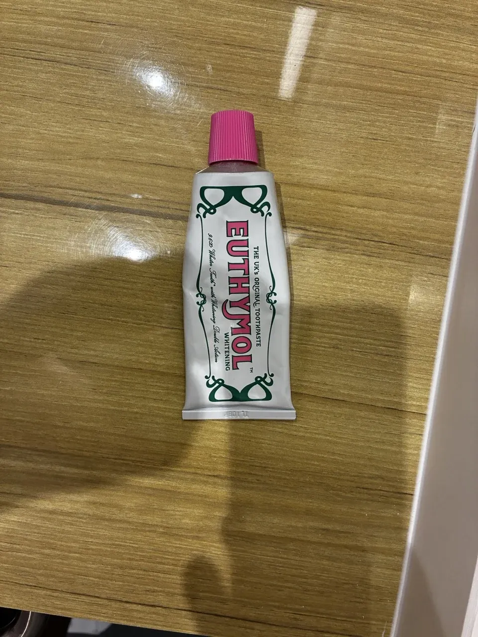 A toothpaste tube with a fluted magenta cap, white body and green ornamental print, photographed on a wooden desk.